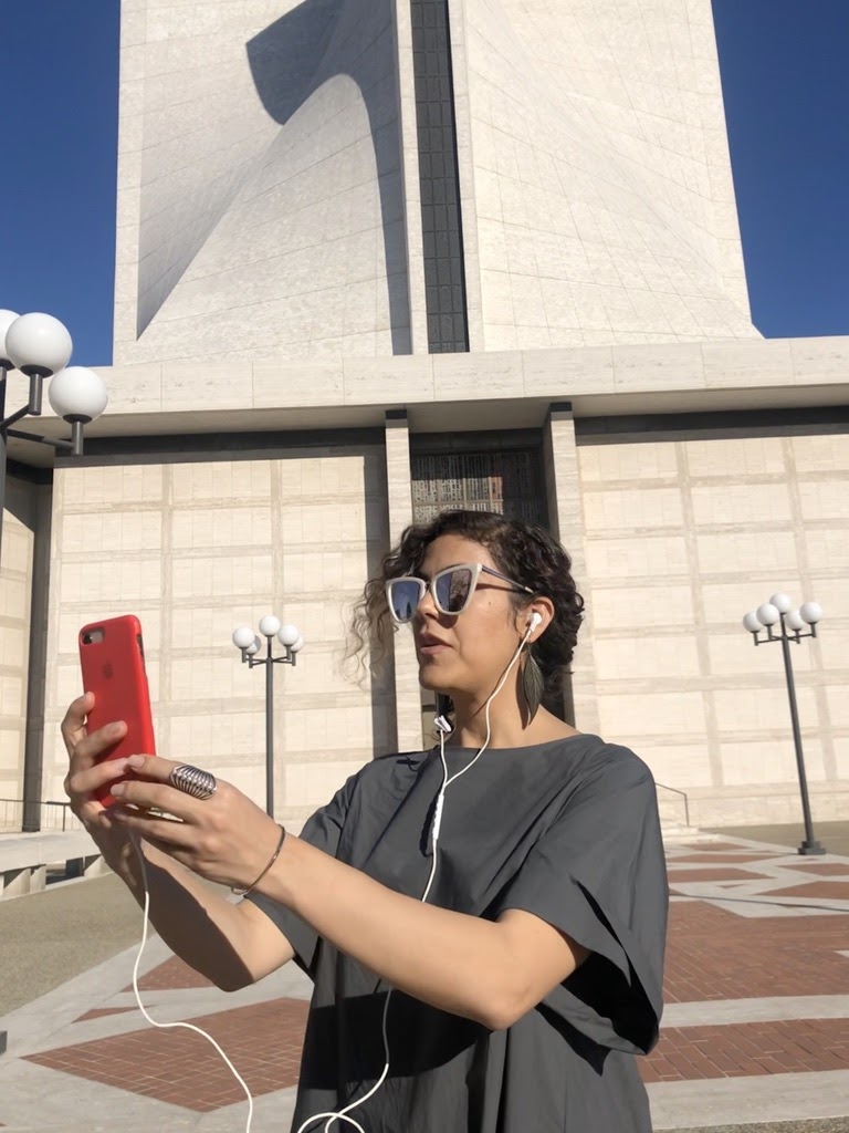 Person standing in front of a building holding up their phone with headphones plugged into it