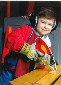 A young child works with a wooden power drill wearing headphones on a piece of wood. 