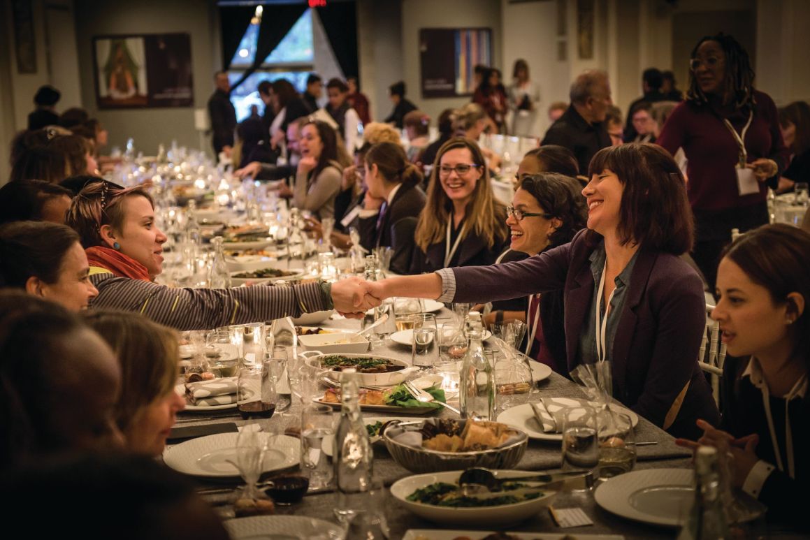 Two women shake hands across a long table where a large group of people sit down for dinner. 