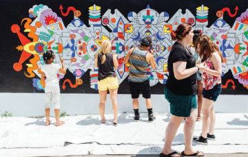 A group of people stand painting an exterior wall with a white woman walking by.