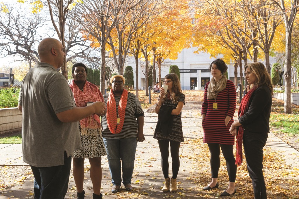 A group of four women stand in a semi-circle listening to a man speak in an outdoor setting.