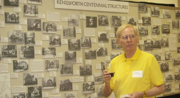 A white man in a yellow collared short sleeve shirt wearing a nametag stands in front and to the right of a wall with the words Kenilworth Centennial Structures and various black and white pictures mounted on it.