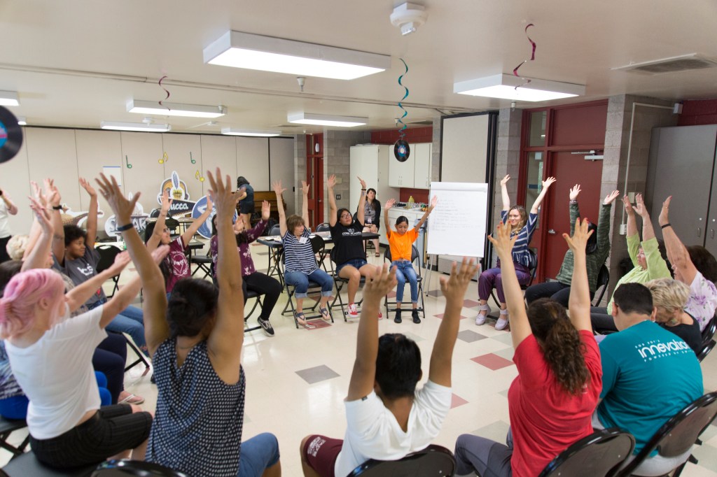 A group of people sitting in a circle all lifting their hands over their heads at the same time.