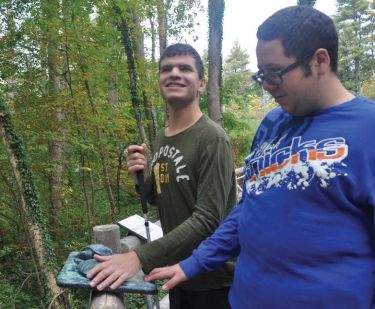 Image of two young people standing in front of a touchable display outside, one appears to be blind and is carrying a walking cane. 