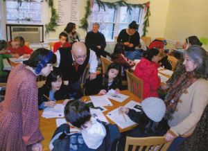A group of young people sit around several wooden tables with adults overseeing a writing project.