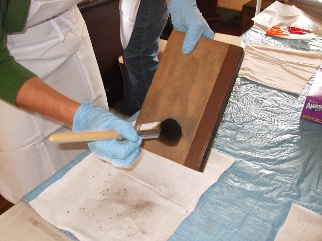 View a pair of plastic gloved hands holding up a book at an angle while dusting with a soft bristle brush. 