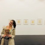 Image of Angela Gala standing in a gallery in front of a series of framed artwork. 