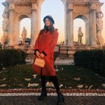 Angela Gala stands in a red coats stands in front of a Roman ruin. 