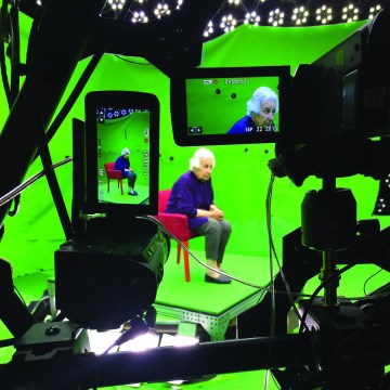 A woman sits in a red chair wearing a blue suit in a green screen theater setting. Several pieces of recording equipment sit in the foreground recording the woman in the chair.