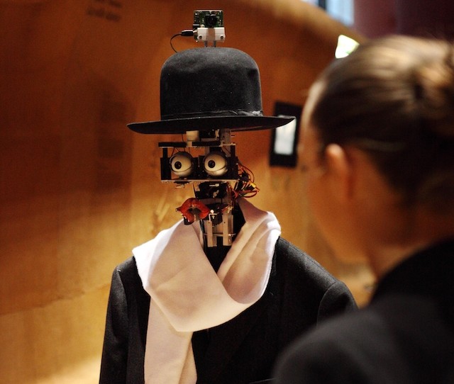 A woman stands speaking with a robot with no face but has eyes an lips wearing a top hat and scarf with a jacket.