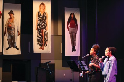Two poets stand at microphones with music stands in front of them.