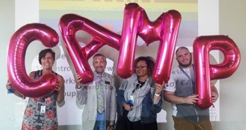 Four members of CAMP are seen holding large, hot pink mylar balloons above their heads, one letter for each person, which spell out CAMP.