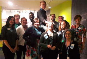 12 members of the CAMP group working to make museums more LGBTQ-friendly pose for a group photo indoors. Some wear badges and name tags. One appears to stand on a ladder or stepstool.