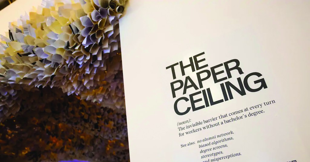 A giant paper flower with white and yellow petals that is attached to a ceiling and wraps around a wall that says, "The Paper Ceiling".