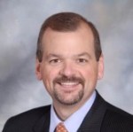 Headshot of a man smiling a thte camera wearing a blue button down shirt and red tie with dark suit.