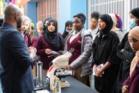 A diverse group of individuals are listening to an employee who is speaking at a table, with a microscope on the table, demonstrating.