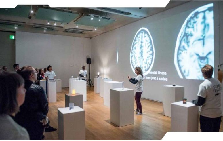 Various people watch a woman presenting in a museum exhibition on homelessness. 