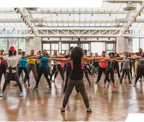 A large group of people stand with their arms outstretched and legs apart with a yoga instructor in front with her back to the camera.