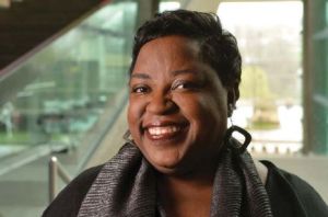 Headshot image of Cecile Shellman, a black woman, standing in front of a geometric shaped stairway atrium area. 
