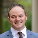 Headshot of Christopher Bedford standing outside wearing a blue suit and brown tie.