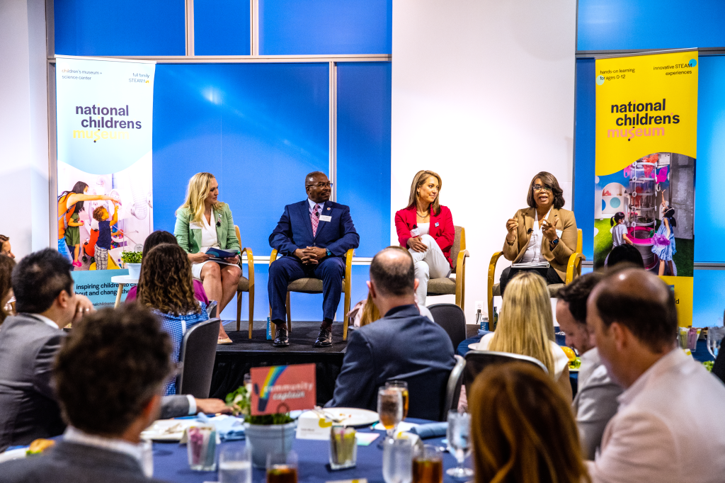 An audience seated at tables in front of a stage watching four speakers on the stage engage in conversation
