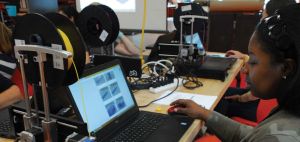 A young black woman sits in front of a laptop computer attached to a string of yellow spool of string.