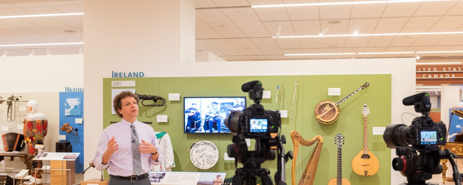 A person speaking in a museum gallery of musical instruments from Ireland in front of cameras