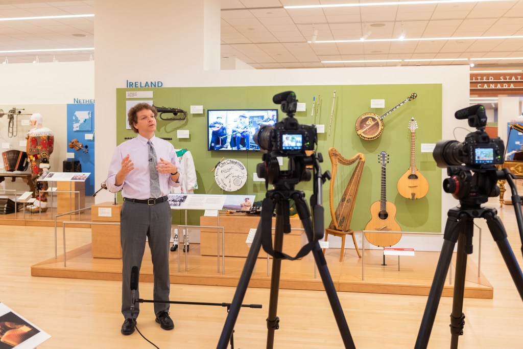 A man stands in front of an exhibit display with several musical instruments facing two video cameras talking about the exhibit.