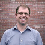 A man stands smiling in front of a brick wall wearing a blue button down shirt