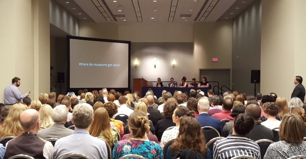 A group of people in an auditorium watching a panel with a screen that reads "Where do museums get data?" behind them.