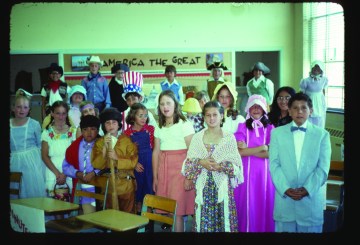 A group of children stand in various costumes behind small wooden desks in a classroom setting.