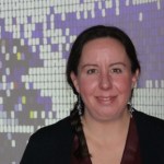 A white woman stands smiling at the camera with her dark brown hair in a ponytail hanging over her right shoulder. She is standing in front of a digital display with small squares in different colors. 