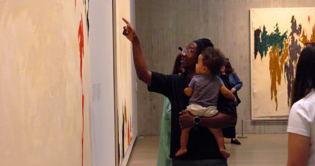 An adult holds a child while pointing at a painting, both looking at the artwork