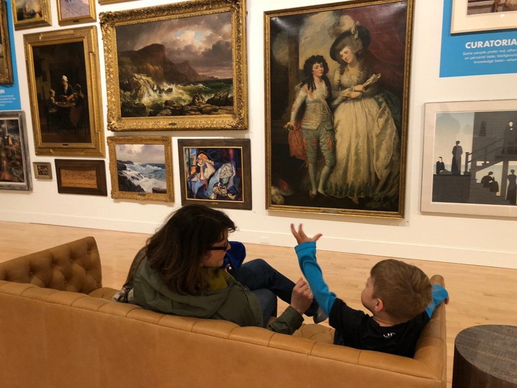 A mother and her son sit on a couch facing a grouping of paintings on the wall opposite. 