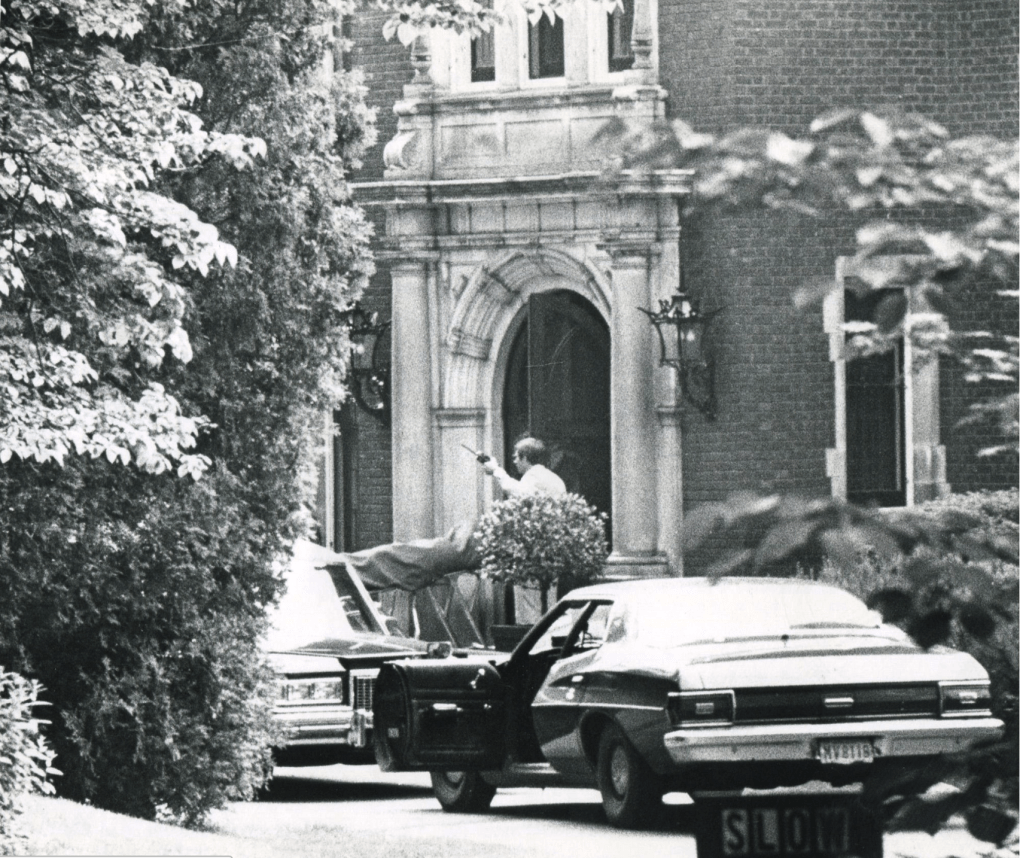 Black and white image of police cars parked in front of the mansion with one gentleman standing near the front door gesturing with what appears to be a walkie talkie in his hand. 