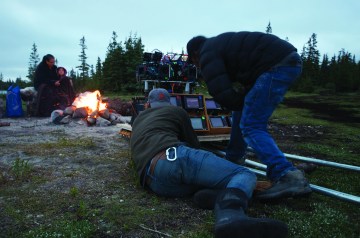 Two people crouch next to film equipment in front of a bonfire in the great outdoors. 