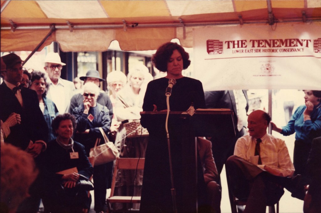 View of a woman standing at a podium with various people in the background.