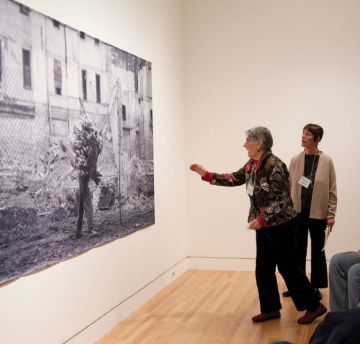 Two women examine a large format photograph on the wall. One of the women is reaching toward the image.