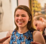 A woman is smiling joyfully while chatting with a few other people. She is wearing a blue lace dress and she has long brown hair. 