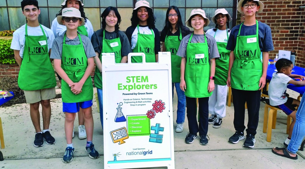 A group of about eight young volunteers stand next to a sign that says STEM Explorers. All of them are wearing bright green aprons.