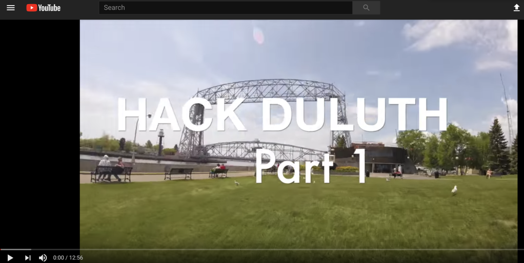Image of a video screen with the words Hack Duluth Part 1 overlayed on top of an image of a bridge and park like area with people and dogs milling about. 