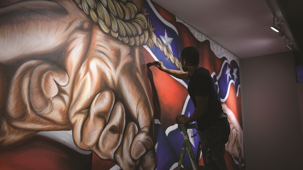 A man stands on a stepstool with a paintbrush in his hand. He is painting a wall mural with hands crossed tied at the wrists overlayed on a Confederate battle flag.