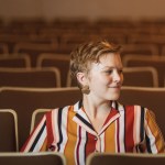 Image of Hannah Hethmon sitting in a theater alone turning her head to the side wearing a striped top. 