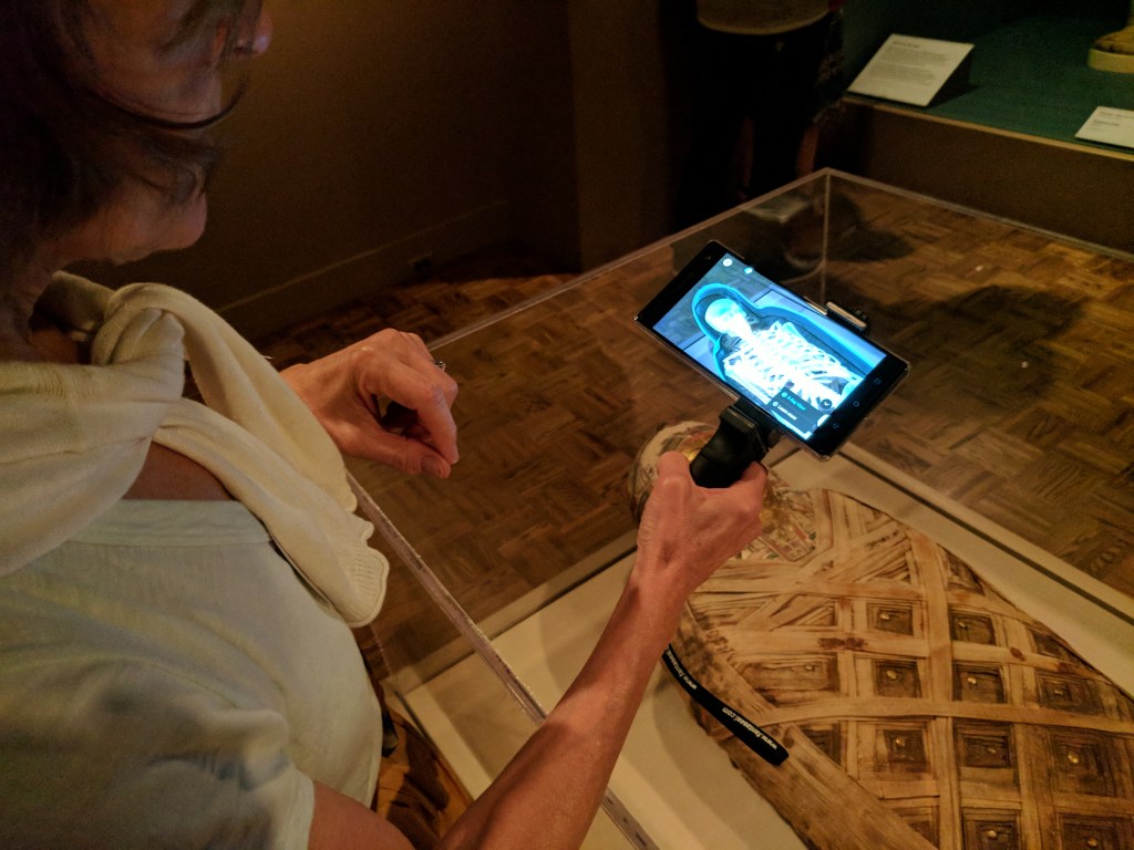 A white woman holds a phone holster in her right hand showing an x-ray of a mummy as she stands at a vitrine with a mummy displayed inside. 