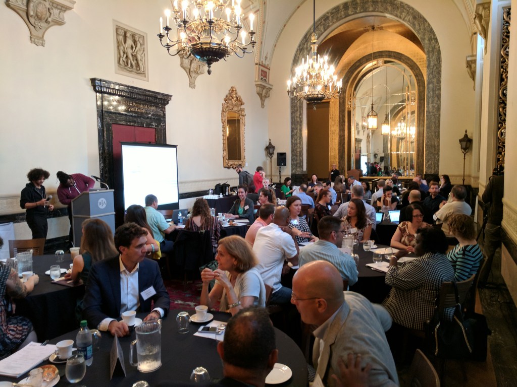 A large group of people sitting around tables sitting in a long ornately decorated hallway. 