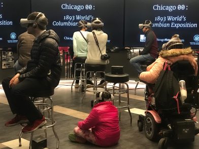 Participants wearing VR headsets in a room with screens reading "Chicago 00: 1893 World's Columbian Exposition"