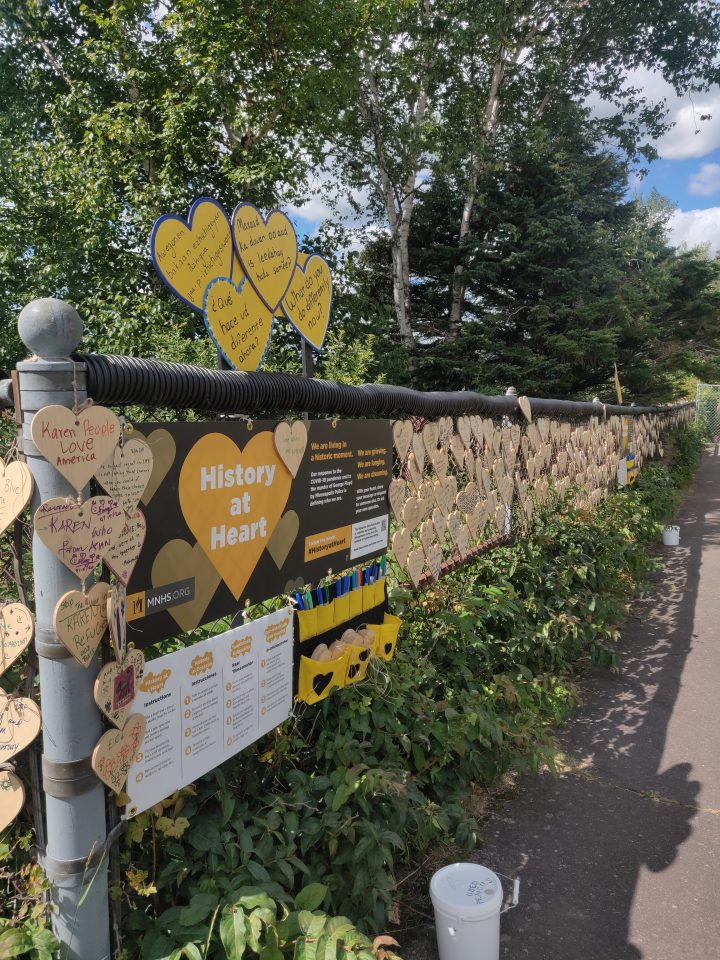 The start of a chainlink fence with a sign reading "History at Heart" and prompts in multiple languages, plus a stock of blank hearts and markers to complete the activity.