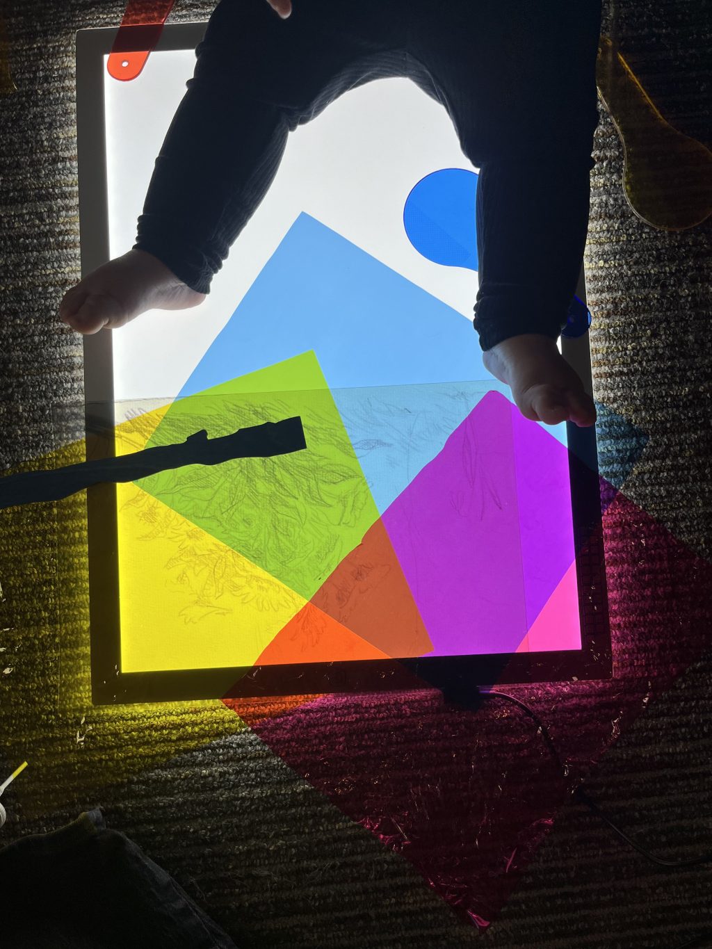 A baby sitting on a light table, with colorful transparent plastic sheets on the surface