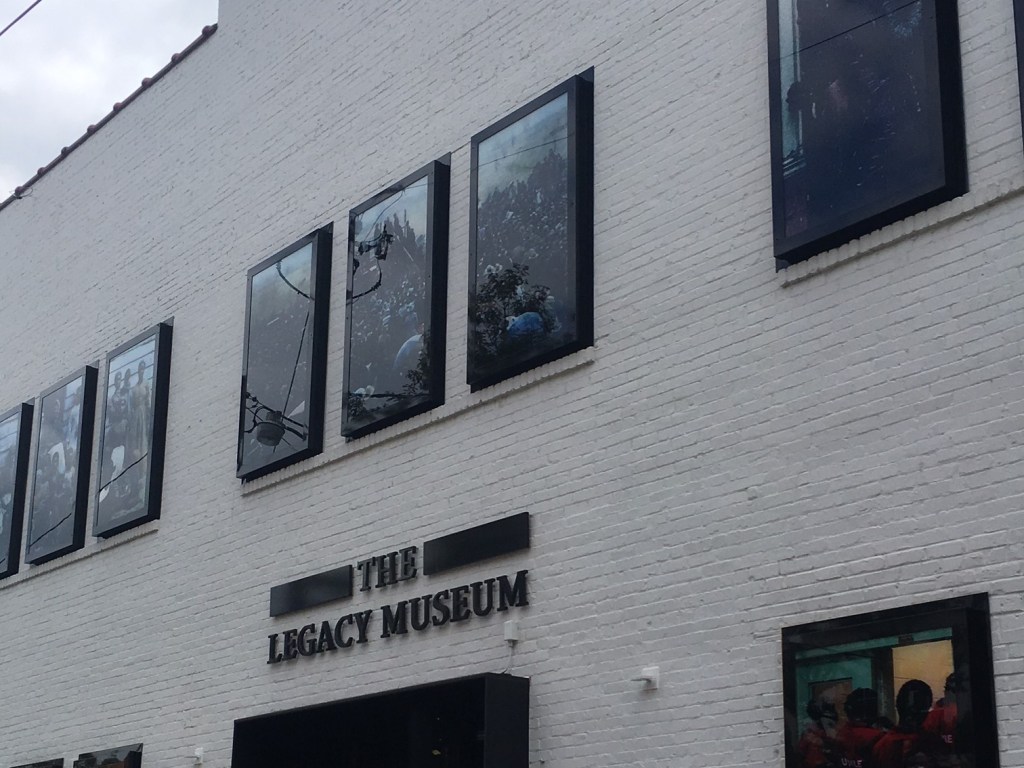 Image of the exterior of the museum with the words "The Legacy Museum" written on the front of a white painted brick building with sets of three dark windows across the front two story building. 