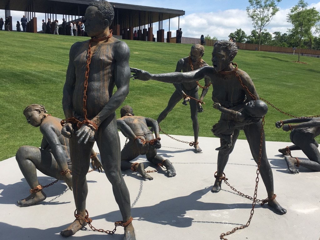 Image of a set of statues at the memorial with several enslaved people chained and reaching for each other. One woman carries a baby as she reaches toward a tall man in chains. 
